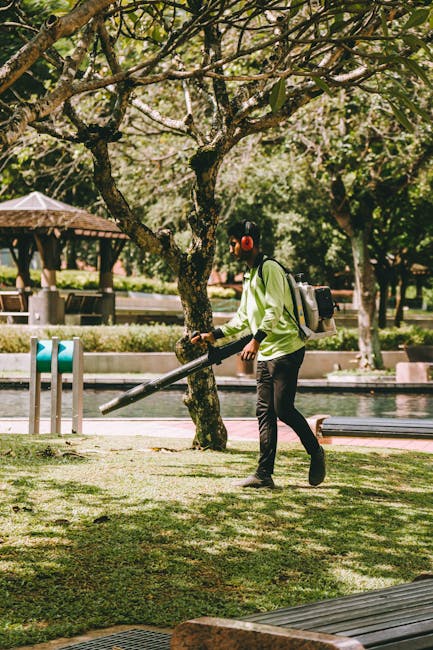 A person wearing a light green jacket, black trousers, and red earmuffs is engaged in garden maintenance, using a leaf blower on a grassy area under a large, leafy tree. The scene features well-kept l